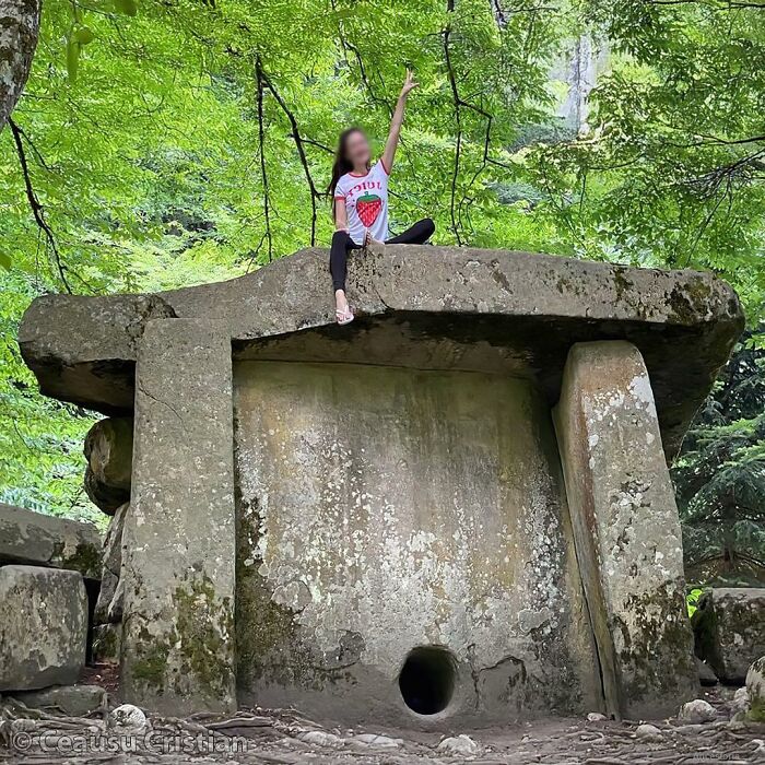 Ancient history stone dolmen monument surrounded by green trees with a person sitting on top in a forest setting
