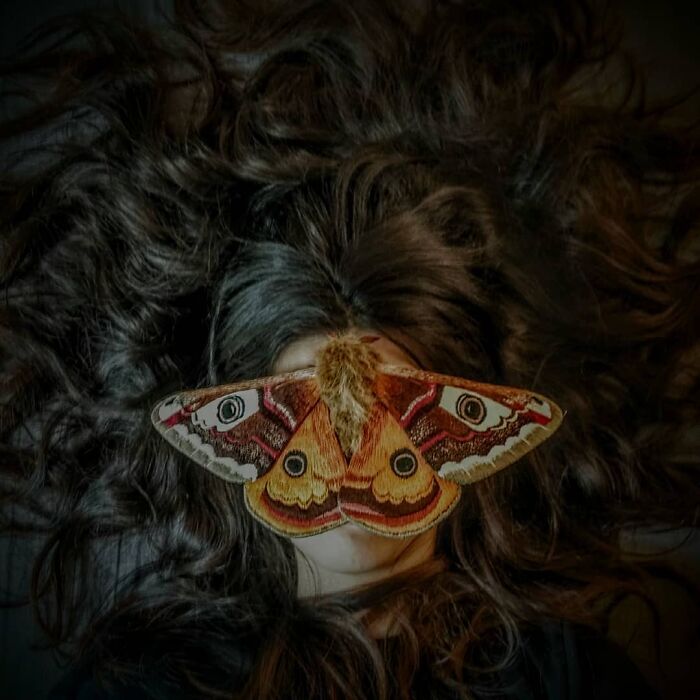 Embroidered fiber moth with detailed colorful wings placed over closed eyes amid dark wavy hair showcasing fiber art.