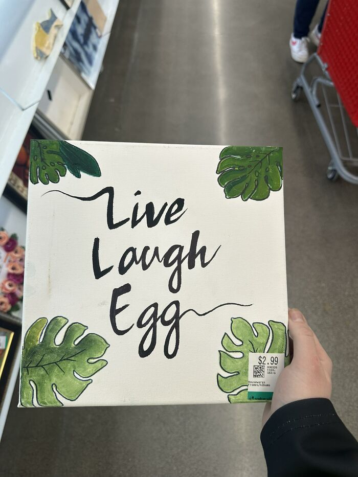 Hand holding a thrifted quirky wall art with phrase Live Laugh Egg surrounded by green leaves for weird wonderful finds.