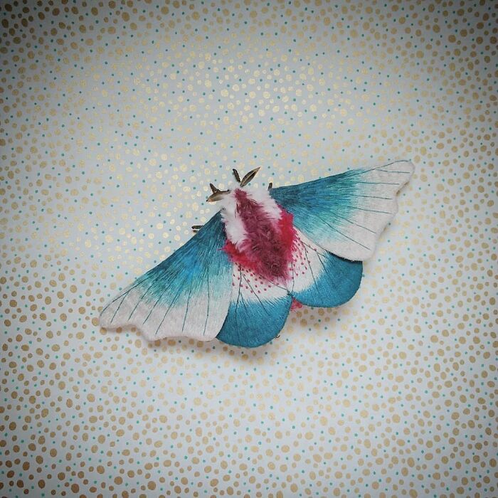 Embroidered fiber moth with blue and pink wings displayed on a dotted beige fabric background.