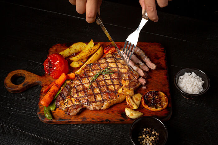 Person slicing a grilled steak with roasted vegetables on a wooden board, illustrating everyday items becoming stupid expensive.