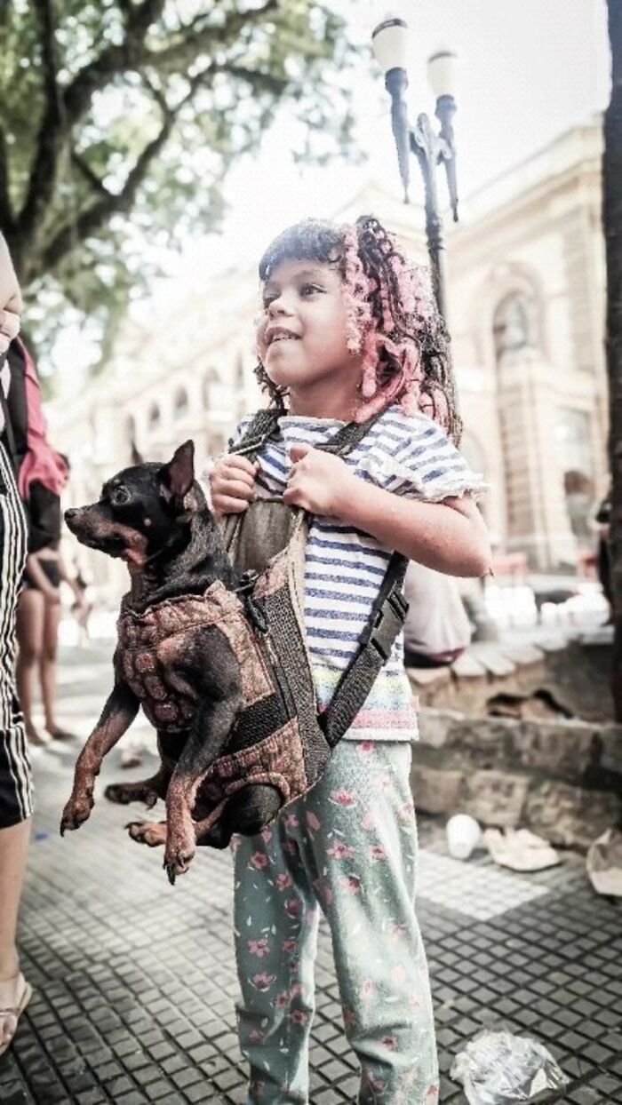 Young child carrying small dog in harness, highlighting the lives of homeless people and their dogs in urban setting.