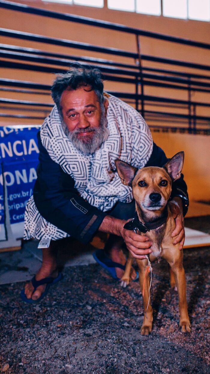 Homeless man wearing a patterned blanket crouching and holding his brown dog in an urban outdoor setting at night