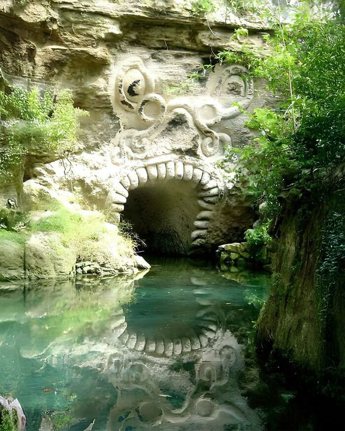 Ancient history stone carving of a mythical face entrance reflected in clear water surrounded by lush greenery.