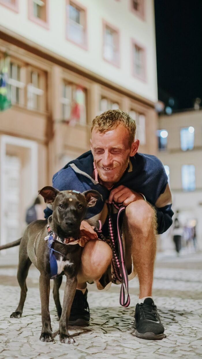 Homeless man smiling and crouching next to his dog on a city street at night documenting lives of homeless people and their dogs.