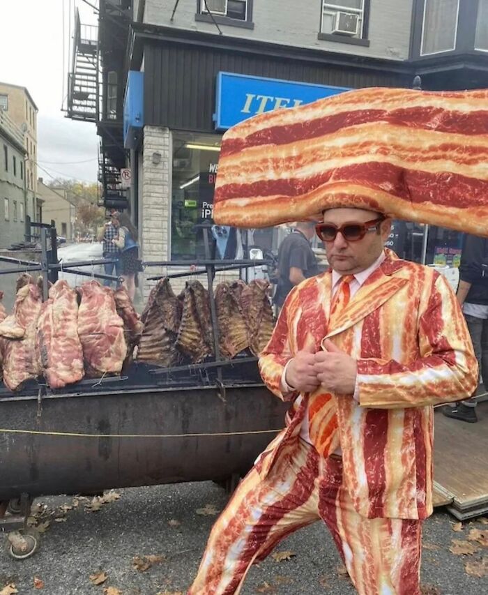 Hombre con traje y sombrero de tocino posando frente a costillas asadas en un asador en la calle, imágenes malditas.
