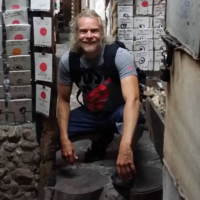 Man smiling and crouching inside a narrow stone and wood corridor filled with stacked boxes, notable people who passed away in 2025.