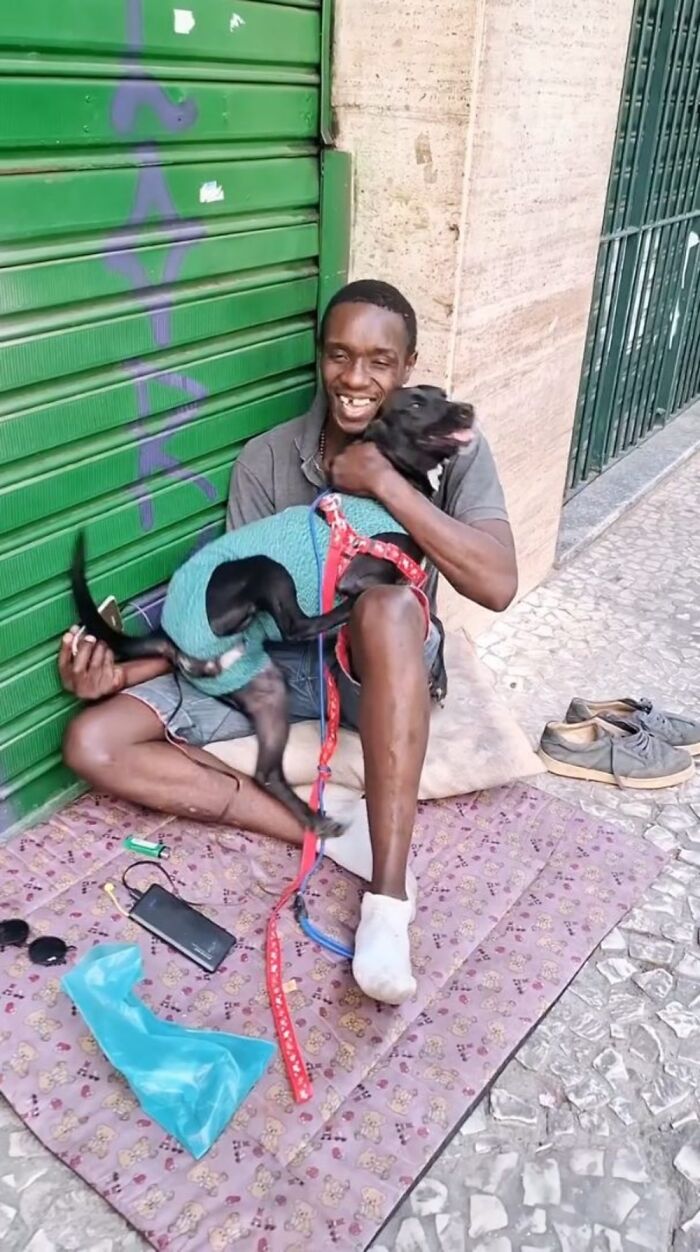 Homeless man sitting on a sidewalk mat, smiling and holding his dog wearing a sweater, showing the bond between homeless people and their dogs.