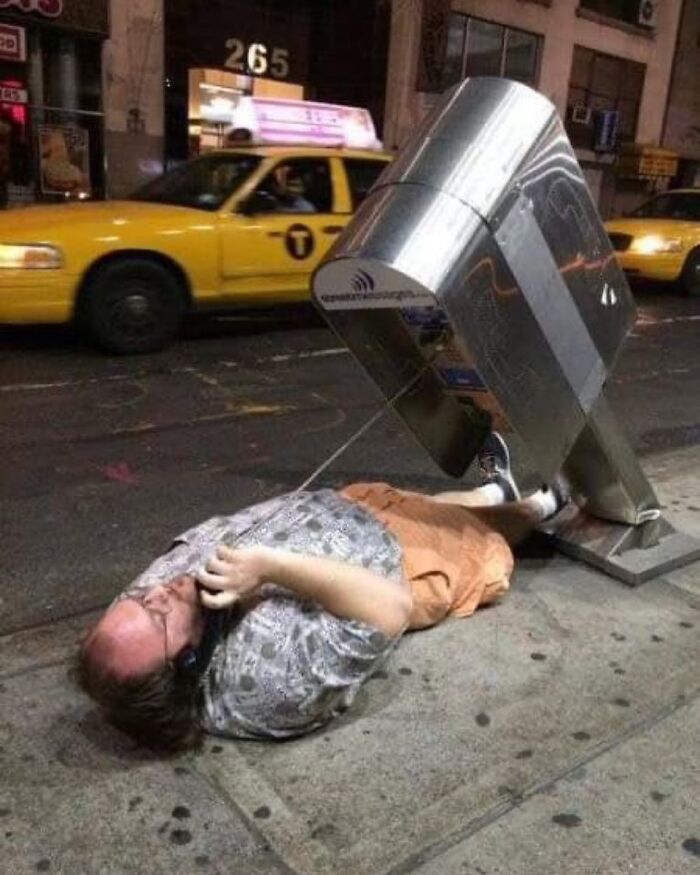 Hombre en situación incómoda al caer junto a un teléfono público, imagen maldita urbana con taxi amarillo de fondo.