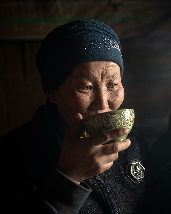Elderly person wearing a blue headscarf, sipping tea from a patterned cup in a softly lit, intimate setting.