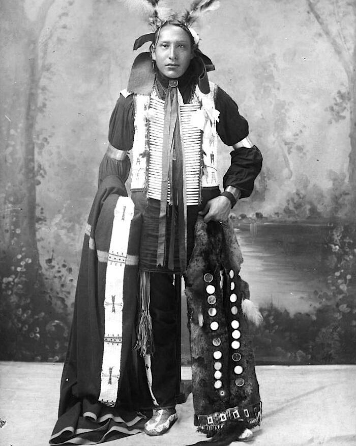 Native American man dressed in traditional attire, posing for a powerful portrait from over a century ago.