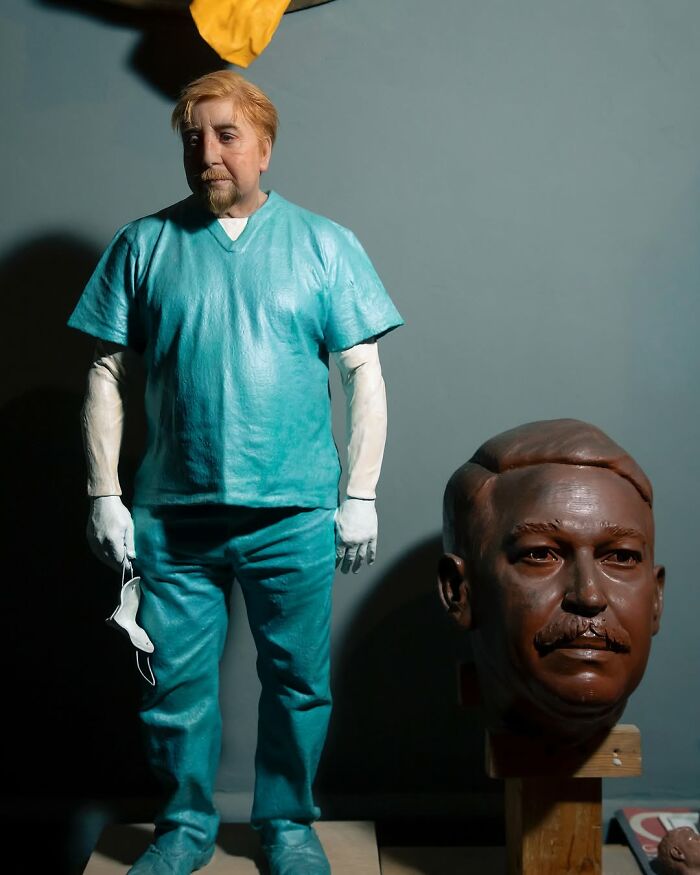 Hyper-realistic sculpture by Ruben Orozco Loza of a man in teal scrubs holding a face mask next to a detailed sculpted head.