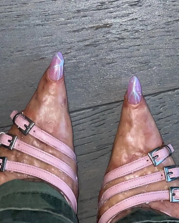 Pies con uñas largas pintadas de rosa y blanco, usando sandalias rosas sobre piso de madera en imagen maldita.