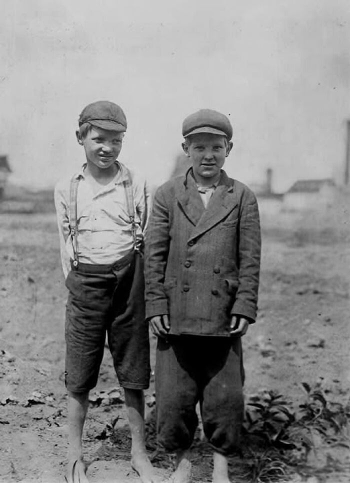 Two young boys barefoot in worn clothes and caps standing outdoors in an antique historical photograph from the past.