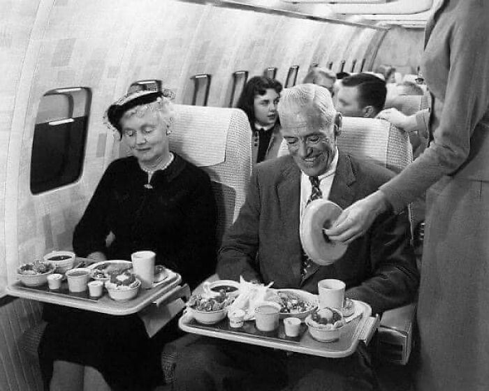 Elderly couple enjoying in-flight meal served by flight attendant in antique historical photographs black and white image.