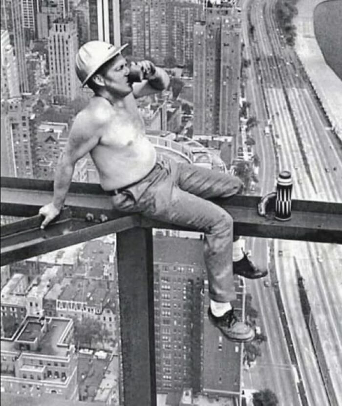 Shirtless construction worker wearing a hard hat drinking from a cup while sitting on steel beam in antique historical photograph.