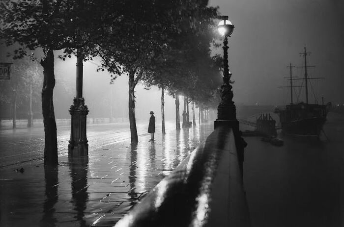 Antique historical photograph of a solitary figure standing by a riverside walkway under street lamps at night.