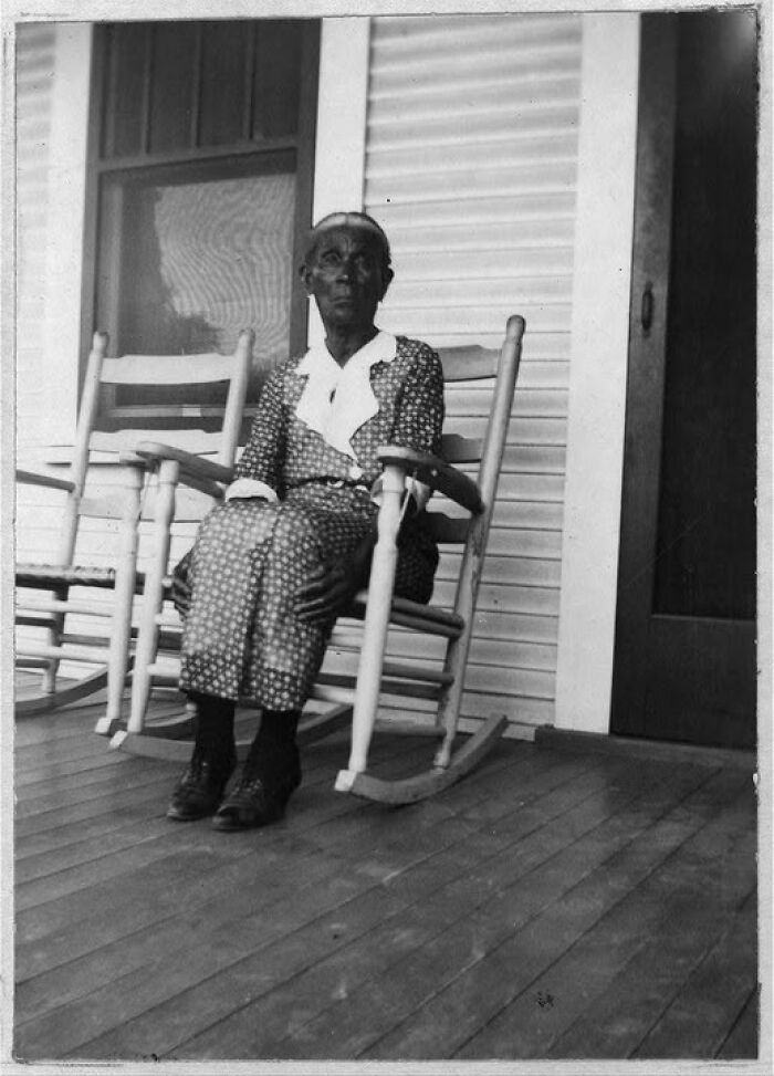 Elderly woman sitting on a wooden rocking chair on the porch in an antique historical photograph.