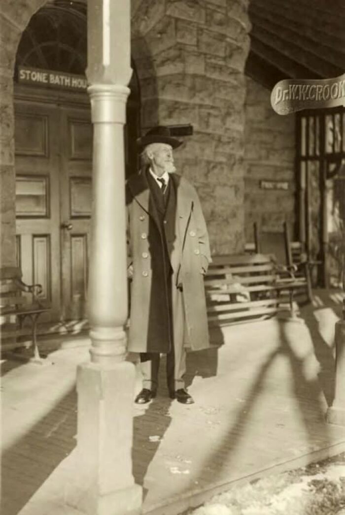 Elderly man in long coat and hat standing outside a stone building in an antique historical photograph.