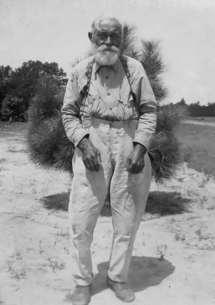 Elderly man with white beard wearing suspenders and loose trousers in an antique historical photograph outdoors.