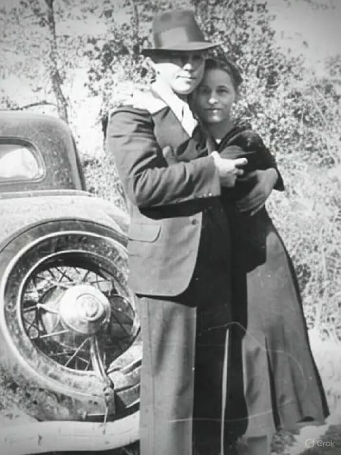 Vintage black and white antique historical photograph of a couple embracing near an old car outdoors.