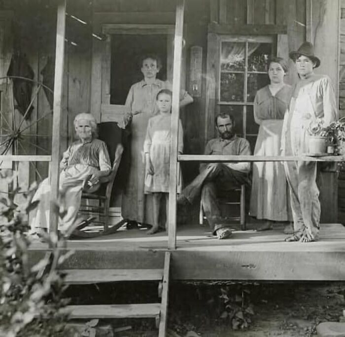 Black and white antique historical photograph showing a family gathered on a wooden porch in old-fashioned clothing.