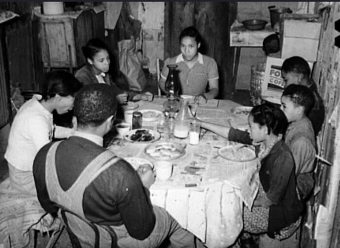 Family sharing a meal around a table in a rustic kitchen, captured in an antique historical photograph.