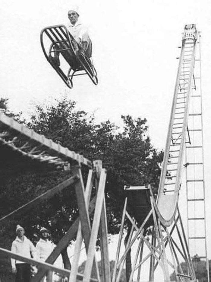 Man flying through the air on a chair launched from a homemade wooden ramp in an antique historical photograph.