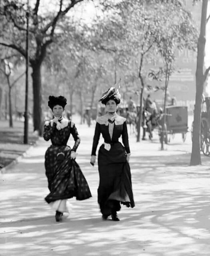 Two women in antique historical photographs walking on a tree-lined path wearing period dresses and hats.