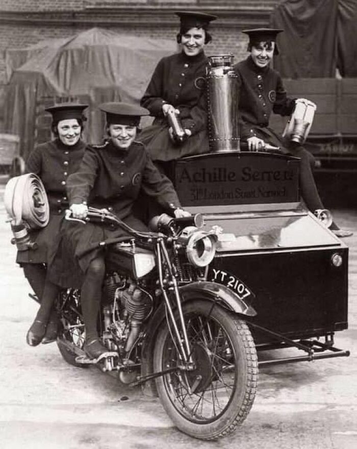 Four women in vintage uniforms posing with an antique historical photograph featuring an early motorcycle and sidecar.
