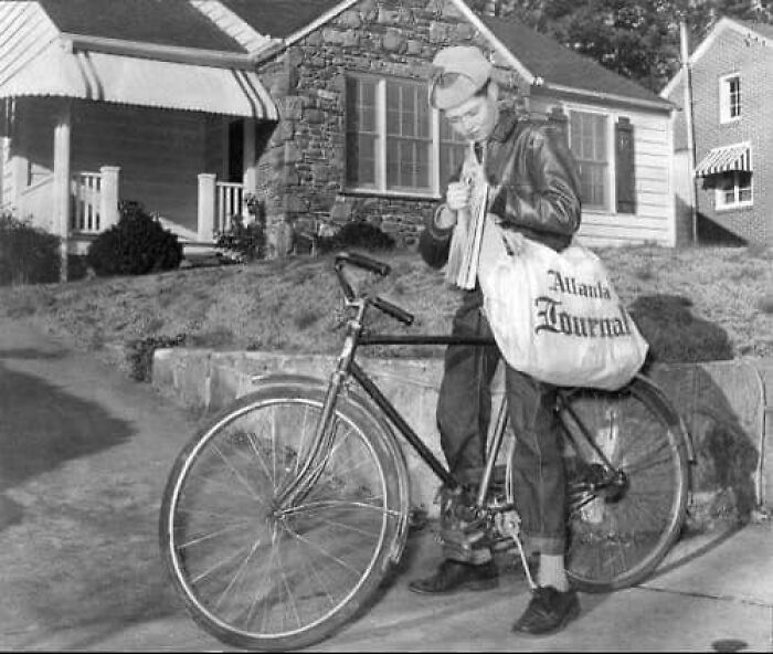 Boy delivering newspapers with bicycle in front of residential homes in an antique historical photograph.