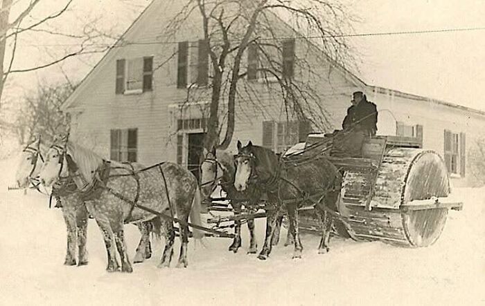 Antique historical photograph of a man driving a horse-drawn roller in a snowy rural setting by a farmhouse.