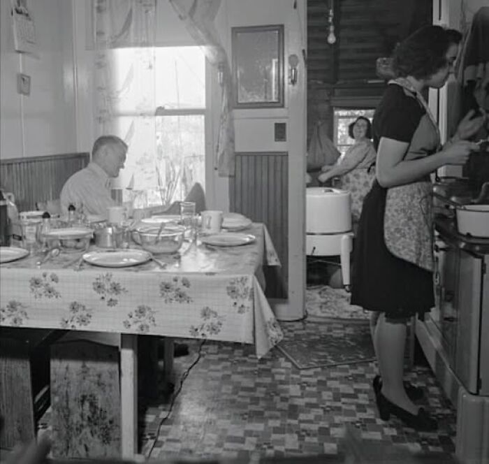 Older black and white antique historical photograph showing a woman cooking and people sitting in a vintage kitchen.