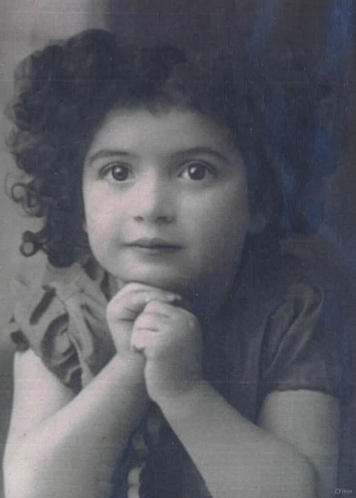 Vintage black and white antique historical photograph of a young girl with curly hair resting her chin on folded hands.