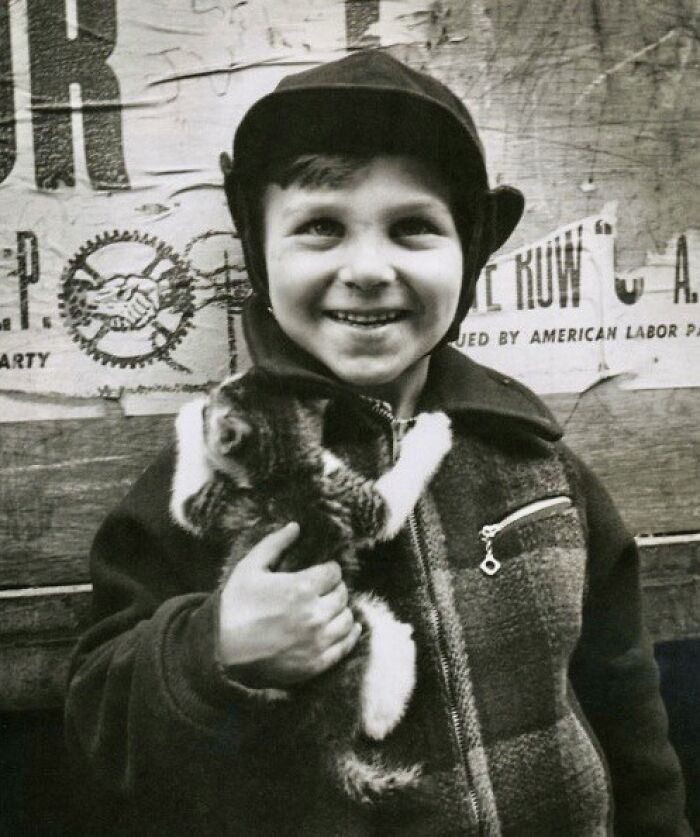 Smiling boy in vintage clothing holding a kitten in front of a weathered poster, an antique historical photograph from the past.