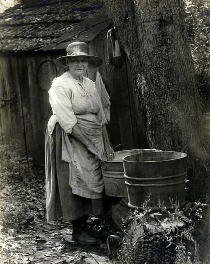 Elderly woman in antique historical photograph washing clothes outdoors by wooden buckets near a tree.