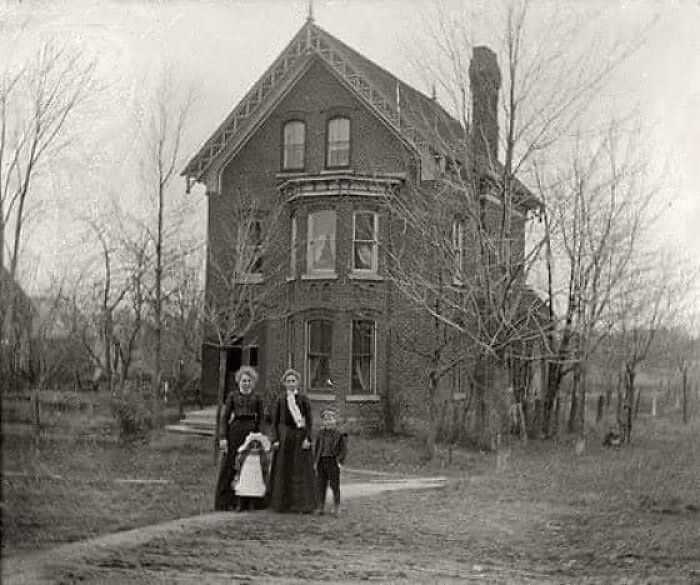 Antique historical photograph showing two women and two children standing outside a large brick house in a leafless wooded area.