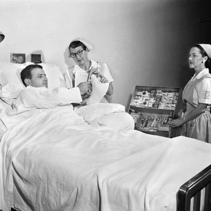 Patient with a broken leg in a hospital bed receiving care from nurses in an antique historical photograph.