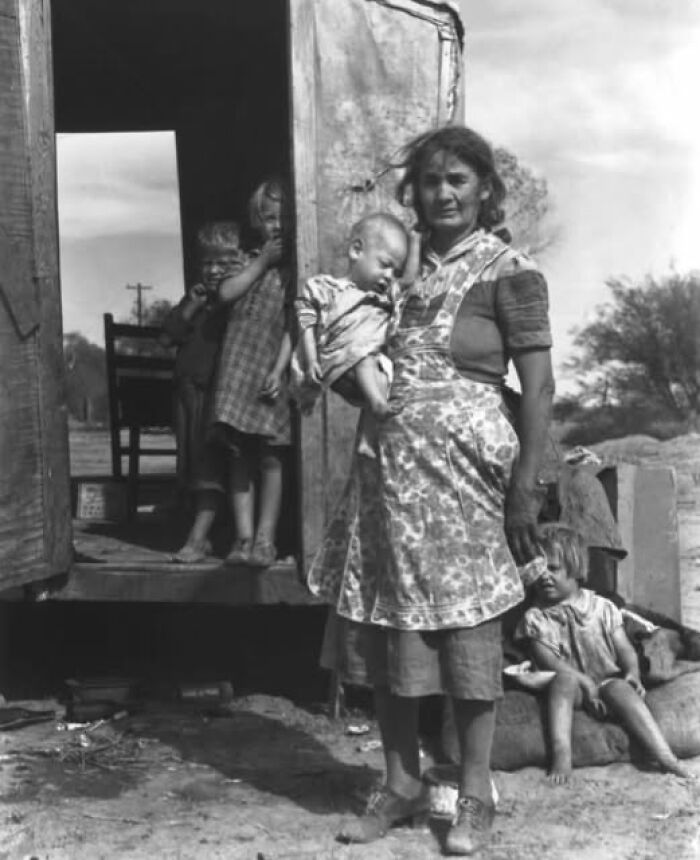 Woman holding a baby outside a rustic home with children nearby in an antique historical photograph from the past.