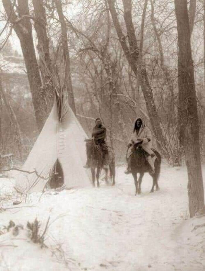 Two Native Americans on horseback near a teepee in snowy woods in an antique historical photograph.