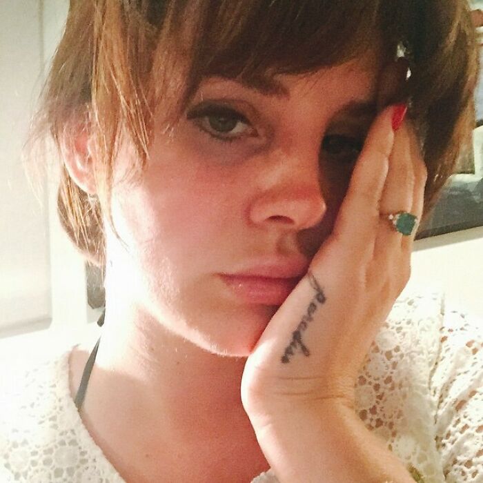 Close-up of a celebrity with short hair resting face on hand showing a tattoo and a green ring in a casual setting.