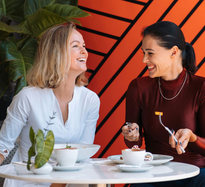 Two women laughing and enjoying food at a table, illustrating problematic behaviors women get a pass for in conversations.