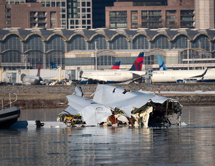 Wreckage of a plane crash partially submerged in water near an airport with Delta planes and buildings in the background