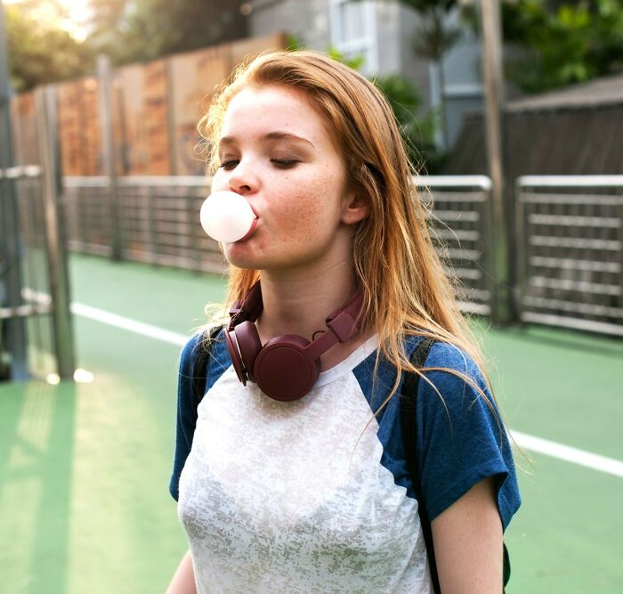 Teenage girl blowing bubblegum bubble outside, wearing headphones around neck, illustrating excessive laziness concept.