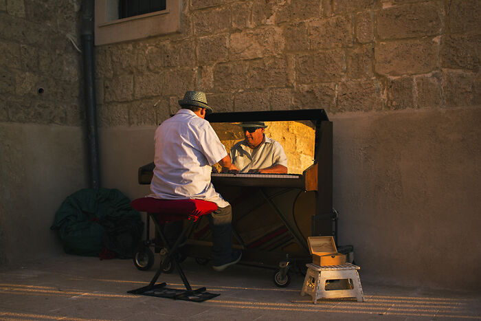 Street photography moment of a man playing piano outdoors near a stone wall, captured by talented photographers.
