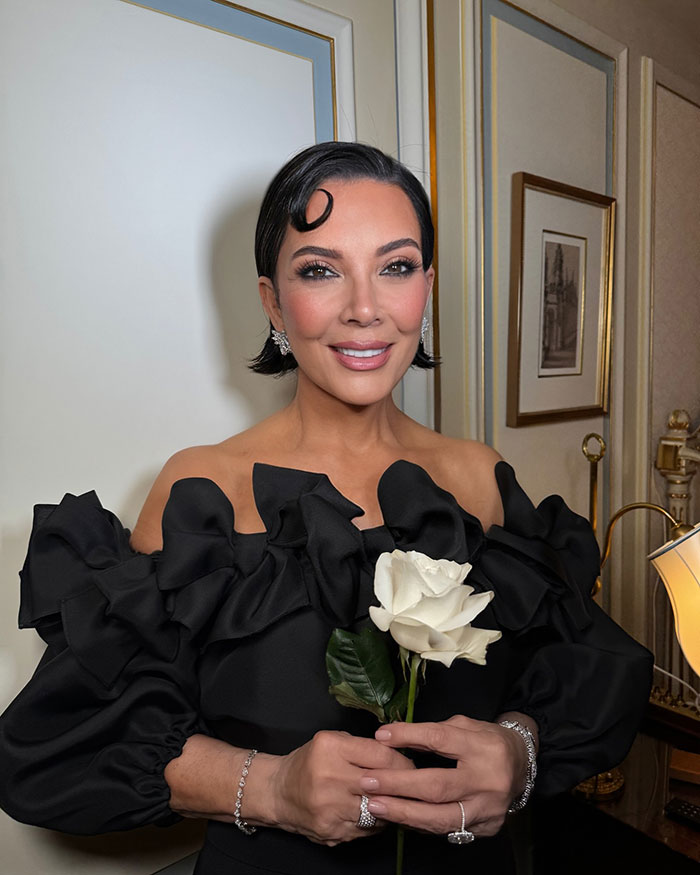 Woman in an elegant black dress holding a white rose, styled with vintage hair and delicate jewelry in a classic room setting.
