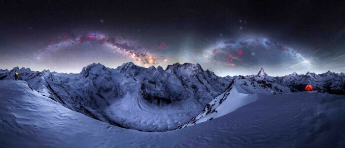 Panoramic night view of the Milky Way arching over snow-covered mountain peaks with a glowing tent in the distance.