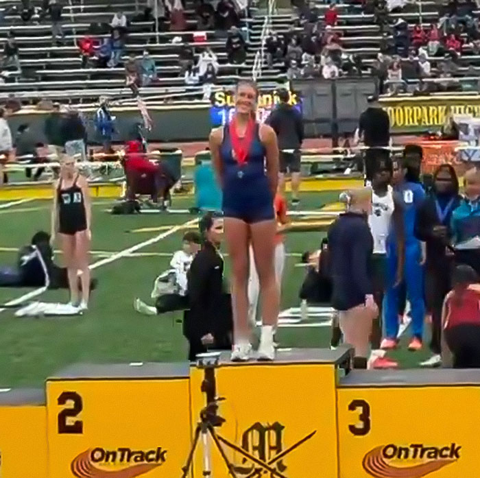 High school girl standing defiantly on first-place podium during track event with competitors in background