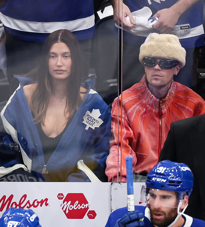 Hailey Bieber watching hockey game in blue jacket beside Justin wearing sunglasses and fur hat in arena seating. Hailey Bieber watching hockey game in blue jacket beside Justin wearing sunglasses and fur hat in arena seating.