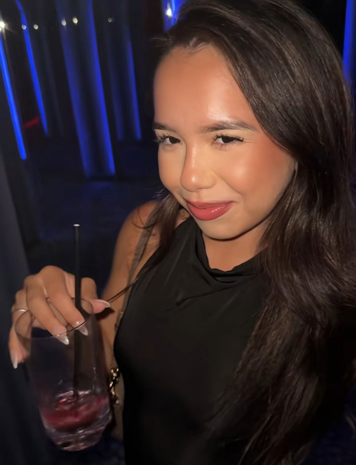 Young woman with long dark hair holding a drink, smiling softly in a dimly lit setting after TSA bag mix-up incident.